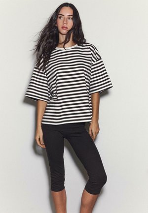 Donna con lunghi capelli scuri che indossa una maglia larga a righe bianco e nere e leggings neri corti, in piedi davanti a uno sfondo semplice.