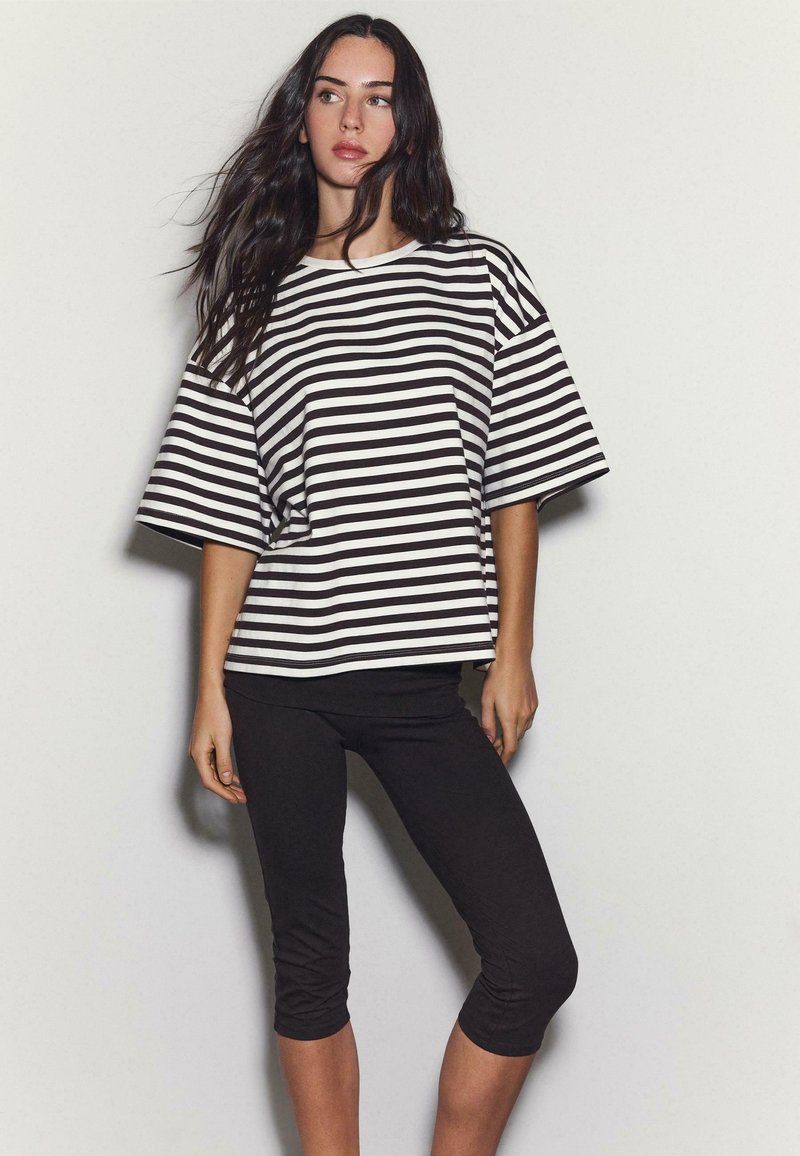 Femme aux longs cheveux foncés portant une chemise ample à rayures noires et blanches et des leggings noirs courts, debout devant un fond uni.