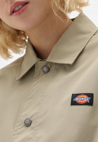 Beige stoffjakke med spiss krage, trykknapper og et rektangulært Dickies-logo-patch på venstre side.