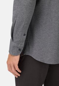 Graues Langarmshirt mit strukturierter Oberfläche, mit einem Knopfdetail am Ärmel und einem abgerundeten Saum. Getragen über schwarzen Hosen.