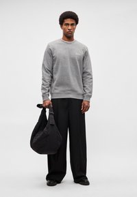 Sweatshirt cinzento com gola redonda e pequena etiqueta, combinada com calças pretas largas. Uma mala preta de ombro macio é segurada na mão esquerda.