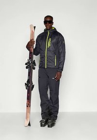 Vyras juodame slidinėjimo džemperyje, kelniuose ir batuose laiko porą slidžių vertikaliai, dėvėdamas didelius juodus slidinėjimo akinius, prieš paprastą foną.