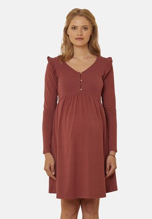 Woman with light brown hair wearing a long-sleeve rust-colored maternity dress with buttons and ruffle details on shoulders.