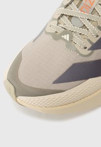 Μπεζ αθλητικό παπούτσι με υφασμάτινη υφή, γεωμετρικές επενδύσεις σε σκούρο γκρι και μαλακή σόλα με πουά σχέδιο.