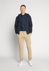Donkerblauwe ritssluiting jack met capuchon, gele logo, over een donkerblauw overhemd en beige oprolbare broek, gecombineerd met witte sneakers.
