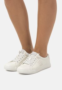 Zapatillas blancas de material de lona, superficie texturizada, diseño de perfil bajo, puntera redonda y cierre de cordones; presenta un sutil logotipo de la marca en el costado.