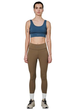 Woman standing facing forward, wearing blue sports bra, brown leggings, white socks, and beige sneakers, with visible arm tattoos on white background.