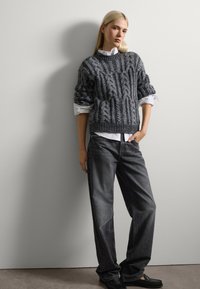 Grijze kabelgebreide trui met opgerolde mouwen, gedragen over een witte colbert. Donkergrijze rechte jeans en zwarte loafers maken de outfit compleet.