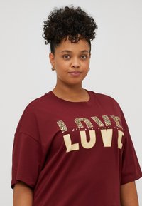 Maroon oversized t-shirt made of cotton, featuring gold and leopard print lettering spelling "LOVE" with additional text below.