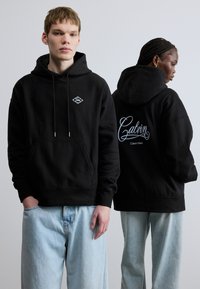 Čierna bavlnená mikina s kapucňou, predným vreckom, šnúrkou na kapucni a nášivkou s logom na hrudi; na chrbte má vyšívaný kurzívny nápis "Calvin Klein".