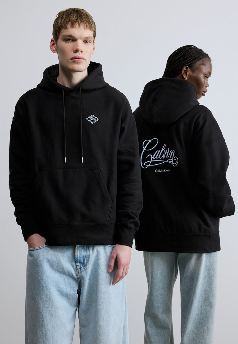 Čierna bavlnená mikina s kapucňou, predným vreckom, šnúrkou na kapucni a nášivkou s logom na hrudi; na chrbte má vyšívaný kurzívny nápis "Calvin Klein".