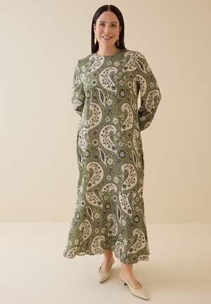 Nainen hymyilee ja hänellä on päällään pitkä oliivinvihreä mekko, jossa on beigejä paisley- ja kukkakuvioita, beiget terävät kengät ja kultaiset korvakorut.