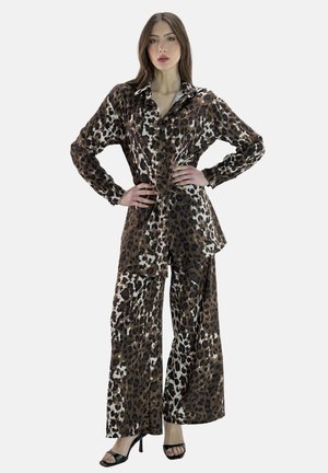 Vrouw die een bijpassende bruine en zwarte blouse met luipaardprint en wijd uitlopende broek draagt, poseert met handen op de heupen, zwarte sandalen met hoge hakken.