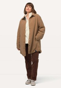 Beige fleecemantel med knäppning fram, sidofickor och krage. Bärs över en ljus skjorta och tillsammans med bruna byxor och grå skor.