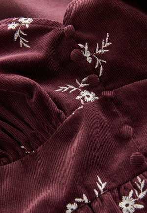 Treisă burgundă din catifea cu broderie albă florală, texturată cu butoane ridicate și detalii pliate. Design moale și detaliat.