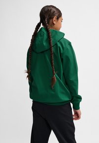 Grön huvtröja med långa ärmar, som har en avslappnad passform och en texturerad tyg. Bakifrån syns flätat hår och en enkel design.