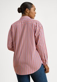 Žena s vlasmi vo vypnutej cope, ktorá má zlatú kruhovú náušnicu a dlhý rukáv s červeno-bielo-modrými vertikálnymi pruhmi, stojaca s mierne otočenými chrbtom.