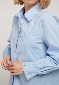 Chemise en coton bleu clair avec fermeture à boutons, une poche ornée d'un détail brodé, et des poignets élastiques avec broderies décoratives.