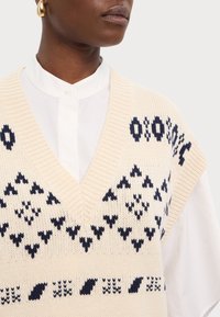 Veste beige en tricot avec un col en V profond, présentant un motif géométrique marine. Portée sur une chemise blanche boutonnée, avec une texture douce.