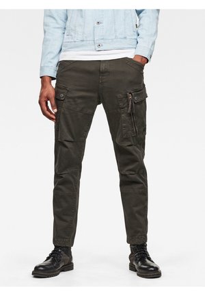 Mężczyzna ubrany w ciemnozielone spodnie typu cargo, czarne skórzane buty, jasnoniebieską kurtkę jeansową, stojący z rękami w kieszeniach.