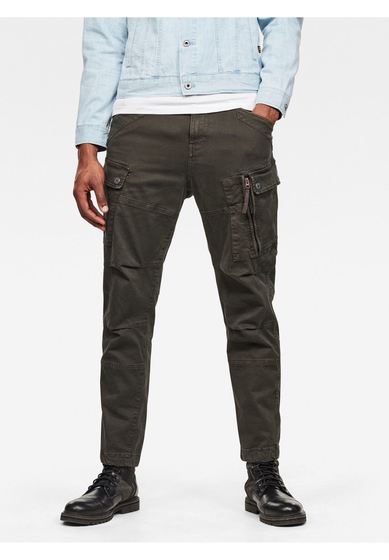 Homme portant un pantalon cargo vert foncé, des bottes en cuir noir, une veste en jean bleu clair, debout avec les mains dans les poches.