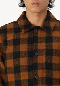 Brun og sort ternet flannel skjorte med krave, frontknapper og en brystlomme. Stoffet ser blødt og tekstureret ud.