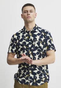 Camisa de manga corta con un patrón abstracto en navy, crema y verde, con cuello en punta y detalle de bolsillo en el frente.