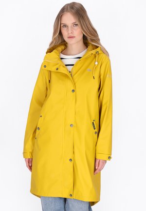 Vrouw met een lange gele regenjas met capuchon, gestreept shirt en blauwe spijkerbroek, staand tegen een witte achtergrond.