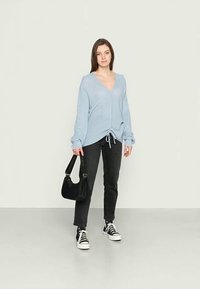 Cardigan de malha azul claro com bainha ajustável, combinado com calças jeans escuras e sapatilhas pretas. Modelo segura uma bolsa preta. Design simples.