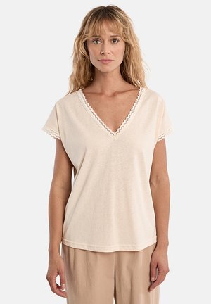 Donna con capelli lunghi biondi che indossa una maglia color crema a scollo a V con bordi smerlati e pantaloni beige, in piedi davanti a uno sfondo semplice.