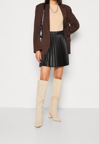 Blazer oversize marrone, top beige, gonna plissettata in ecopelle nera, stivali al ginocchio in suede beige con tacco a blocco e zip laterali.