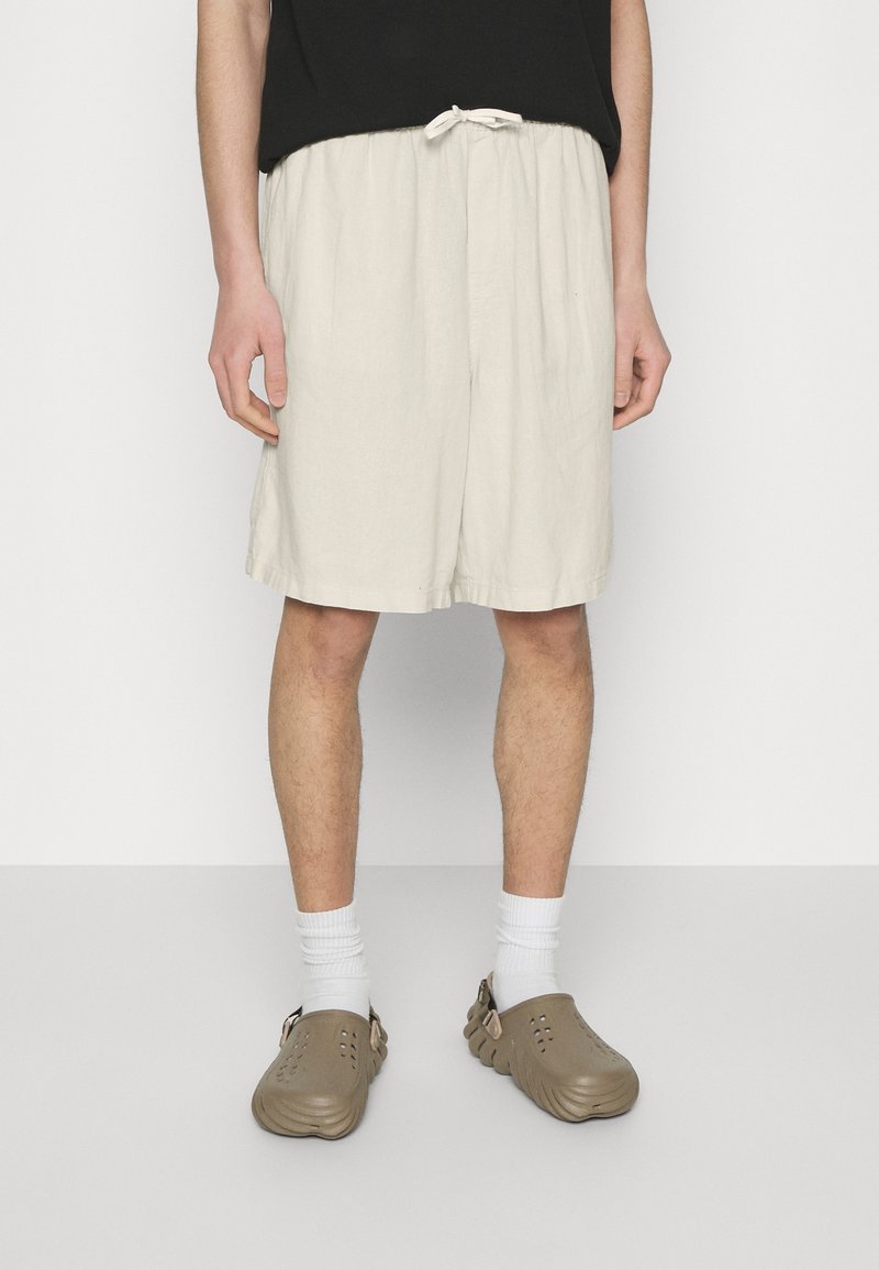 Homme portant un short beige mi-long, des chaussettes blanches et des chaussures marron sans lacets, debout devant un fond blanc uni.