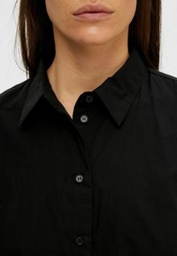 Svart skjorta med knapp-up som har en klassisk krage, slät tygtextur och svarta knappar längs framplacket.