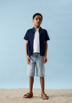 Camicia button-up a maniche corte navy sopra una t-shirt bianca, abbinata a pantaloni shorts in denim azzurro chiaro e sandali marroni. Sfondo sabbioso chiaro.