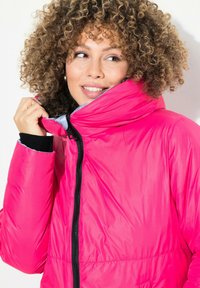 Chaqueta aislante rosa con cuello alto, cierre de cremallera en la parte delantera y forro gris en contraste. Cuenta con puños elásticos y un acabado liso y brillante.