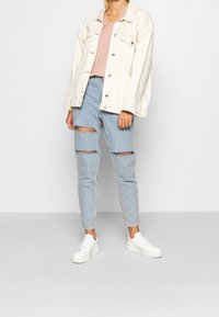 Hellblaue Jeans mit Knie-Rissen, kombiniert mit einem sanften rosa Oberteil und einer übergroßen, cremefarbenen Jeansjacke; weiße Sneaker runden den Look ab.