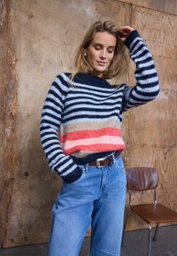 Pull tricot rayé avec des couleurs bleu marine, bleu clair et corail, doté d'un col rond et d'épaules tombantes, accompagné d'un jean en denim bleu clair.