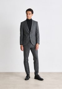 Completo in lana grigia con blazer aderente e pantaloni coordinati, abbinato a un dolcevita nero. Dettagli notevoli: tessuto testurizzato e scarpe nere lucide.