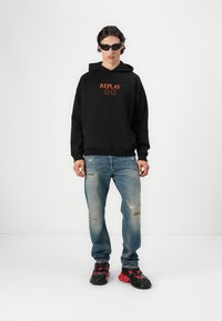 Czarna bluza z kapturem z pomarańczowym logiem "REPLAY", podarte niebieskie dżinsy i czarno-czerwone masywne sneakersy. Dodatki to czarne okulary przeciwsłoneczne.