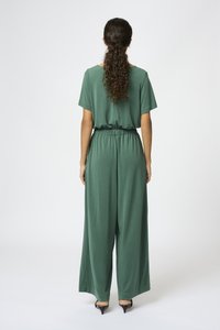 Jumpsuit verde hecho de tela suave, con un corte holgado, mangas cortas, cinturilla elástica y diseño de pierna ancha. Llevado con tacones negros.