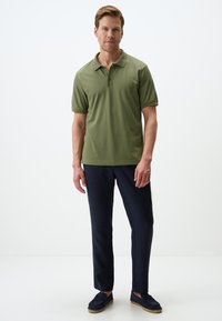 Polo verde oliva con colletto e tre bottoni, abbinato a pantaloni blu navy e mocassini scuri, in piedi su uno sfondo bianco.
