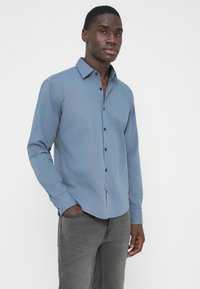 Light blue long-sleeve shirt with a button-down collar, black buttons, and a red accent tag. Slim fit with a smooth texture.