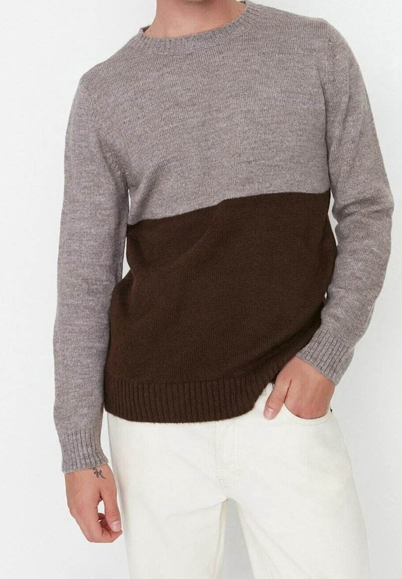 Uomo che indossa un maglione lavorato a maglia bicolore con la parte superiore grigia e quella inferiore marrone, abbinato a pantaloni beige chiaro, mano in tasca, vista ritagliata.