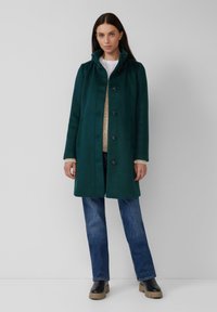 Grüner Wollmantel mit hohem Kragen, Knopfverschluss und Seitentaschen. Kombiniert mit einem beigen Strickpullover und blauer Jeans. Schwarze Blockabsatzstiefel.