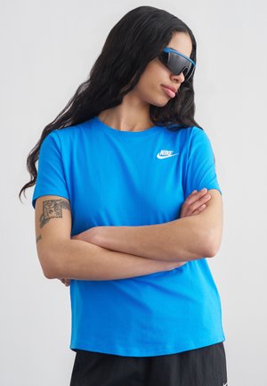 Γυναίκα με μακριά σκούρα μαλλιά φοράει μπλε μπλουζάκι Nike, μαύρα σορτς, μεγάλα φουτουριστικά γυαλιά ηλίου, σταυρωμένα χέρια, τατουάζ στο αριστερό μπράτσο, κοιτάει πλάγια.