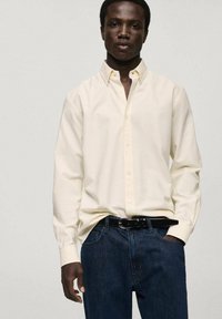 Long-sleeve, light yellow button-up shirt with subtle vertical stripes, paired with dark blue denim jeans and a black belt.