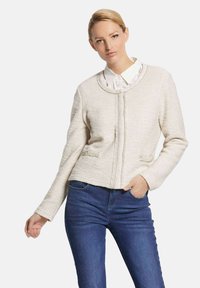 Helle beige gesteppte Jacke mit rundem Ausschnitt, Frontreißverschluss und zwei Taschenakzenten. Über einem weißen Hemd mit Kragen getragen, kombiniert mit blauen Jeans.