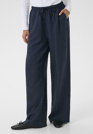 Pantalon large bleu marine en tissu texturé, avec une taille élastique dotée d'un cordon de serrage et des poches latérales. Chaussures plates noires incluses.