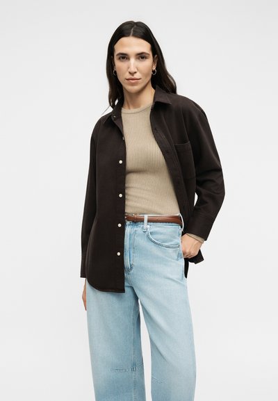 Γυναίκα με μακριά σκούρα μαλλιά που φοράει μαύρο πουκάμισο με κουμπιά πάνω από μπεζ ριμπ πλεκτό τοπ, ανοιχτό μπλε τζιν και καφέ ζώνη, στέκεται μπροστά σε λευκό φόντο.