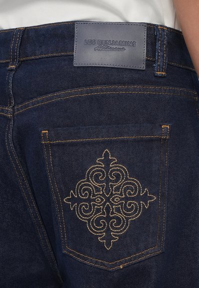 Los jeans de mezclilla oscura presentan un bordado decorativo dorado en el bolsillo trasero, con una etiqueta de cuero grabada en la cintura.